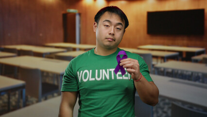 Man in green volunteer shirt holding purple ribbon stands in empty classroom with wooden walls and tables, showing support for a cause and awareness.