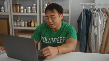 Young man in volunteer shirt using laptop in charity room filled with donation items and supplies.