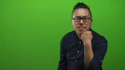 Fototapeta premium Chinese young man wearing glasses and a dark shirt clenches fist and grimaces in green studio; anger focus resolve.