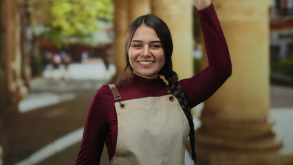 Young woman with braided hair, wearing an apron, dances joyfully in an outdoor street setting with stone columns, conveying liveliness and hispanic culture.
