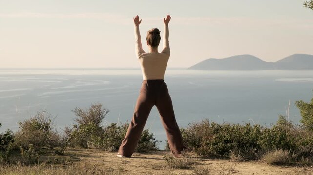 Woman practicing yoga outdoors with panoramic sea view. Full body shot of female doing balance and stretching exercise. Healthy lifestyle, mindfulness, fitness and wellness in nature.