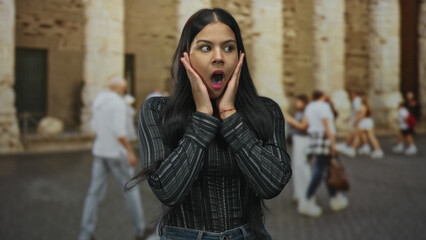 Woman with hands on cheeks and open mouth on street past aged stone columns wearing striped shirt...
