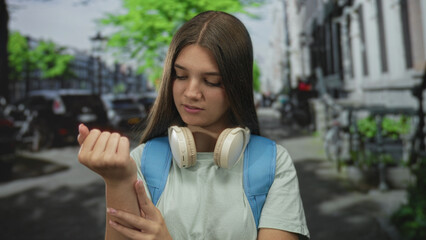 Teen girl with light green shirt and headphones holds bare wrist while inspecting it on street;...