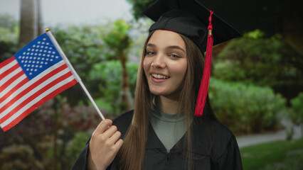 Smiling woman in graduation cap and gown holds american flag in green park; pride achievement celebration success.