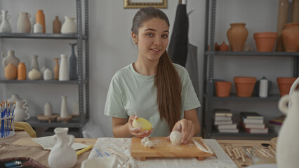 Teenage girl kneads clay with both hands in studio surrounded by ceramic vessels; creative focus mindful practice.