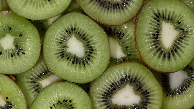 Vibrant seamless loop video of fresh sliced kiwi fruit rotating on a dark surface, filmed from a top-down angle. The dynamic motion highlights the kiwi's bright green flesh, black seeds, and fuzzy bro