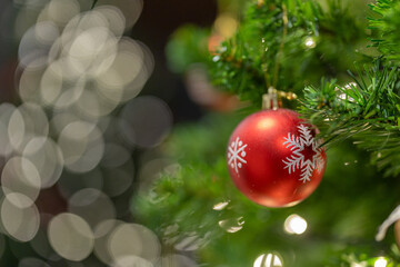 A red Christmas bauble with white snowflake patterns hangs on a green fir branch, softly lit by warm fairy lights, with creamy bokeh in the background creating a calm, festive holiday mood.