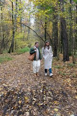 Fototapeta premium Two women walk together along a forest path covered with autumn leaves, tall trees around, natural daylight, relaxed and friendly atmosphere