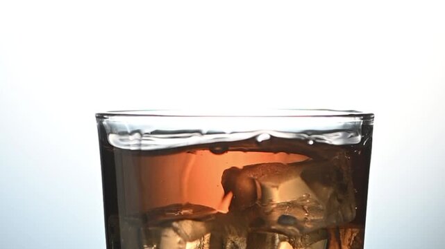 Cinematic slow-motion close-up of whiskey being poured directly onto a large ice cube in a crystal glass. The clip captures the beautiful interaction between the amber spirit and the ice &mdash; the initial