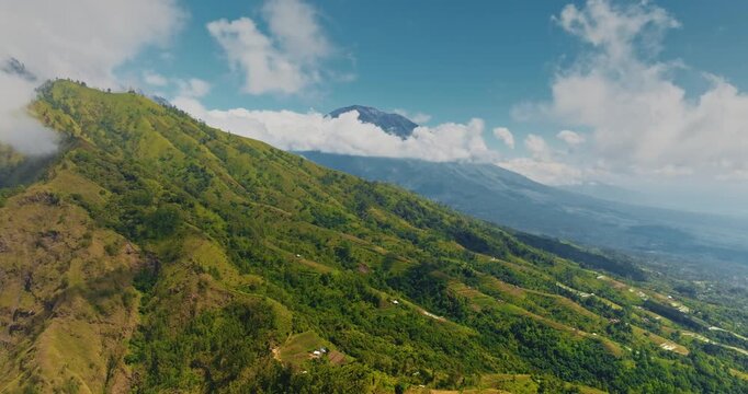 Indonesia, Bali: Lush green slopes of Mount Abang with low clouds and distant Mount Batur rising under a bright blue sky, a dramatic tropical volcanic landscape. Aerial drone flight zoom out panorama