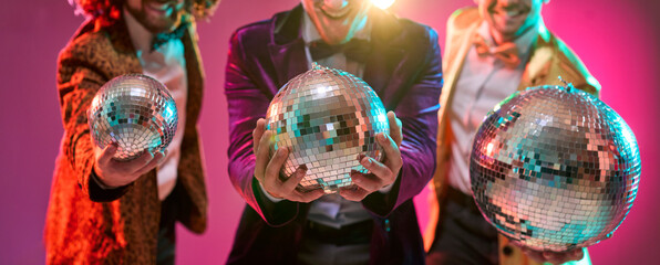 Men dancers party at disco mirrorball celebration. Friends in retro costumes hold sparkling spheres under neon lights, posing for fun party ads marketing. Clear message of joyful celebration.