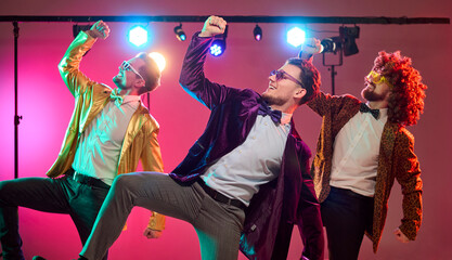 Men dancers in party costumes under lights celebrating. Three friends perform moves on a lit stage as bright lighting creates vibe for music promotion. Concept shows group celebration mood.