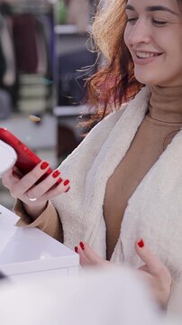 Smiling Woman Making Contactless Payment with Smartphone in Fashion Store. A cashier holds a POS terminal to pay for goods
