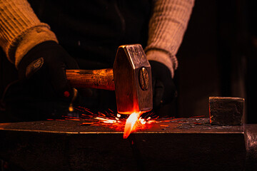 Blacksmith forging hot steel on anvil with hammer, glowing metal and sparks flying in dark workshop, close up hands in protective gloves, traditional metalworking craftsmanship and indus
