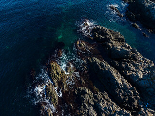 Waves crash against rugged rocks along a serene coastline under a clear blue sky at sunrise