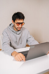 Vertical portrait of young man wearing stylish blue light blocking glasses and hoodie, sitting at desk and typing on laptop, concentrating on screen while working remotely or studying indoors.