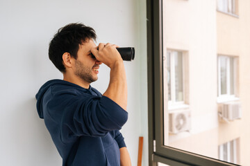 Curious young man looking through binoculars from bright apartment window, actively observing the adjacent building and city environment, engaging in surveillance or neighborhood watch.
