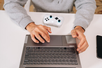 Top view of gamer hands navigating laptop touchpad, using contemporary gaming controller, engaging with digital technology for online entertainment, cloud gaming, web browsing or work.