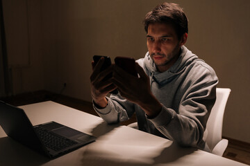 Young man holding two smartphones, focused on comparing apps and messages, multitasking between screens for work or personal digital communication and productivity at home.