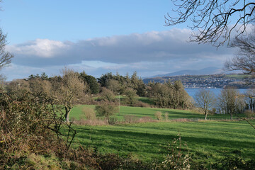 Rolling green field leads to trees bordering a lake, distant mountains under a blue sky with scattered clouds, bathed in bright, natural light in Isle of Anglesey - Wales - UK