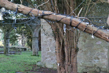 A branch wrapped with lights crosses the frame, leading to a stone archway in a grassy, overcast graveyard, evoking a somber mood in Isle of Anglesey - Wales - UK