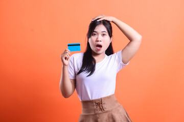 A young woman looks shocked and surprised holding a credit card in hand, looking at camera with mouth open. isolated on orange background