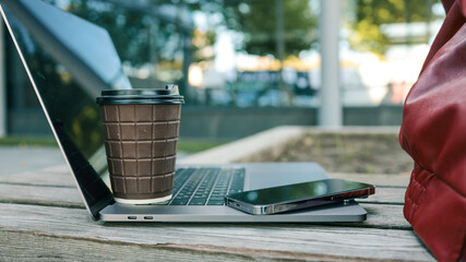 Laptop, coffee cup, and smartphone on a wooden bench outdoors, ready for work or study