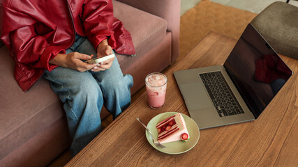 Young person in red jacket using phone while enjoying dessert and laptop at cafe