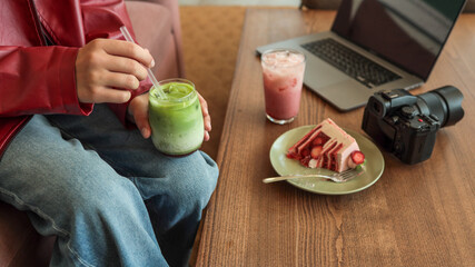 Person holding a matcha latte with a slice of strawberry cake and camera on a wooden table