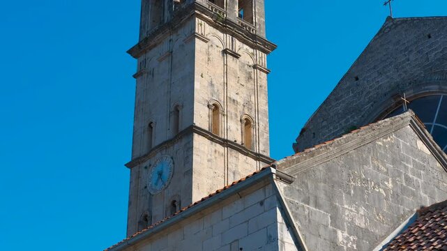 Stone bell tower of st nicholas church in perast montenegro. Upward tilting view of the historic st. Nicholas church bell tower, revealing its ancient stone architecture and clock against a blue sky