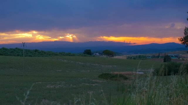 Dramatic sunset with sunbeams over a green countryside landscape. Beautiful golden sunrays breaking through dark clouds during a vibrant sunset over the rolling green hills of a rural landscape