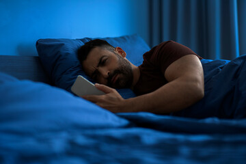 Sleepless man lying in dark bedroom with cellphone in hand and scrolling on social media, guy having insomnia at night