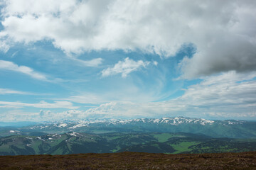 Awesome sunlit top view to forest hills and big snowy mountain range under lush clouds in changeable weather. Scenic hilly alpine landscape with sunlight and shadows. Partly cloudy in high mountains.