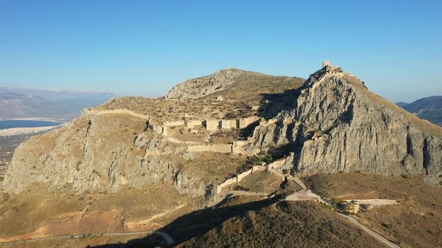 Aerial view of the historic Acrocorinth fortress perched on a rugged hilltop under clear blue skies in Greece. The ancient stone walls and dramatic landscape create a striking scene.