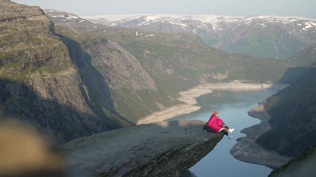 Girl wrapped in sleeping bag is sitting on the edge of cliff on Trolltunga, Norway. Aerial of Norwegian Troll Tongue.