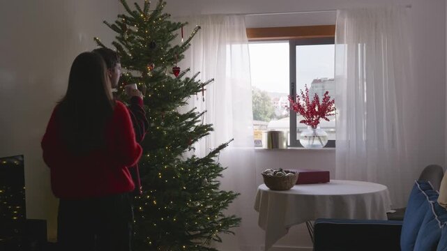 Young couple drinking coffee, hugging while looking at decorated Christmas tree