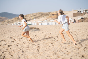 Cheerful girls in casuals running on sandy beach enjoying summer vacation with mountains and blue sky in background