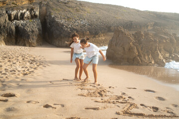Playful girls drawing on sandy beach near sea enjoying fun vacation with rocks and mountains in background during sunny day