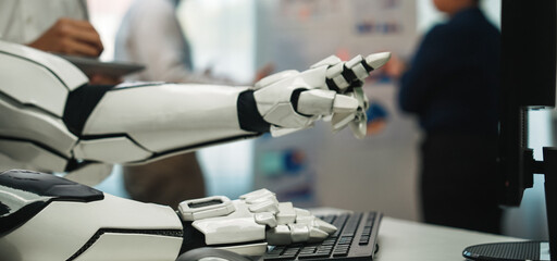 A robotic hand extends towards a keyboard in a modern office. Professionals engage in discussions, highlighting innovation and technology in a collaborative setting.Noogenesis