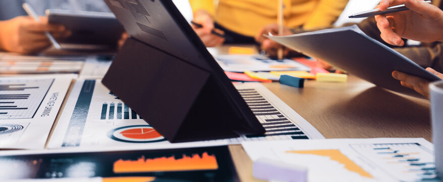 A vibrant workspace captures a diverse team engaged in a business meeting. Professionals collaborate using digital devices and paper reports to analyze data and strategize. SACTR - Powered by Adobe