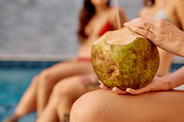 Woman holding a refreshing coconut drink with a straw, relaxing by a pool on a sunny tropical...