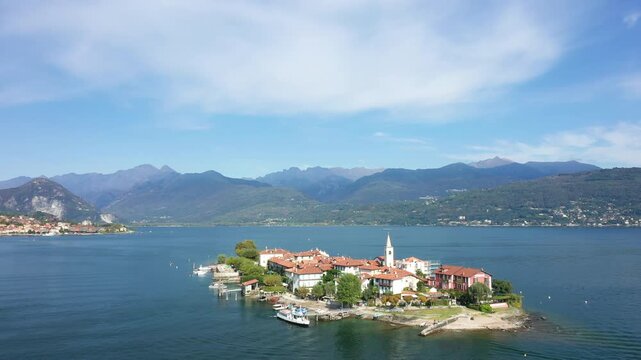 Scenic aerial view of Isola dei Pescatori with traditional buildings, a church, and boats on Lake Maggiore, surrounded by mountains under a blue sky.