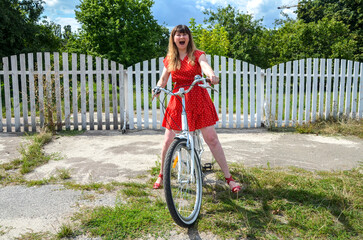 Smiling woman in a red dress rides a bicycle beside a white picket fence on a bright sunny day, conveying playful energy, freedom, and carefree outdoor vibes