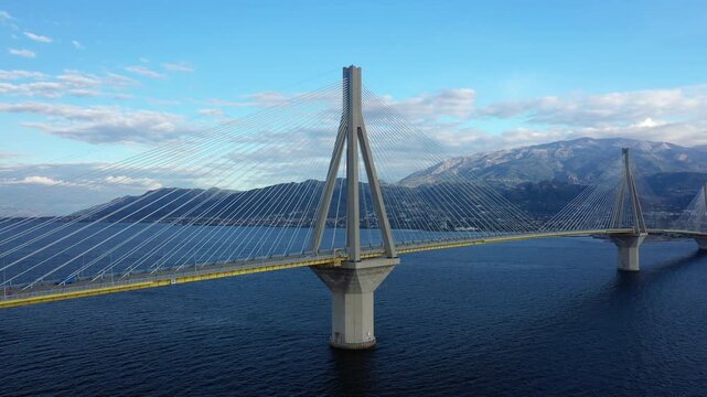 Wide view of the Rion Antirion Bridge spanning across deep blue water with mountains in the background, under a partly cloudy sky in Greece.