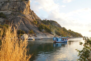 Fototapeta premium A fishing boat going out to sea to fish.A colorful wooden boat sets out to sea at dawn to fish. It's morning, dawn, and hot. In the background is a mountain with green trees and grass and a blue sky.