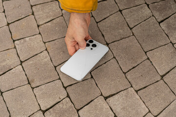 A person lifts a smartphone with a cracked screen over a cobblestone surface outside