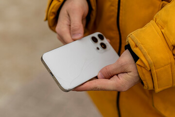 A person stands outside, holding a phone with a cracked screen in one hand, wearing a yellow coat