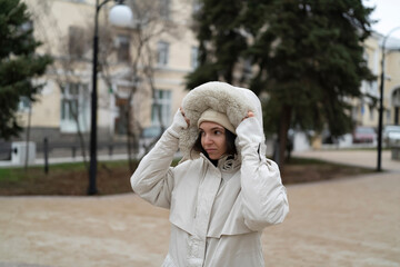 Woman stands in a park and adjusts her fur hat while wearing a winter coat