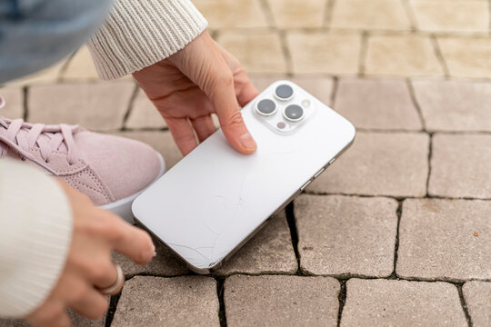 A person reaches down to grab a smartphone that fell on the ground in daylight