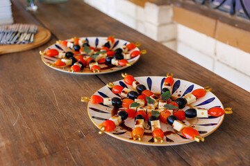 Mediterranean appetizer skewers with cherry tomatoes, olives and cheese served on decorative plates along a rustic wooden table. Catering setup for events and hospitality.
